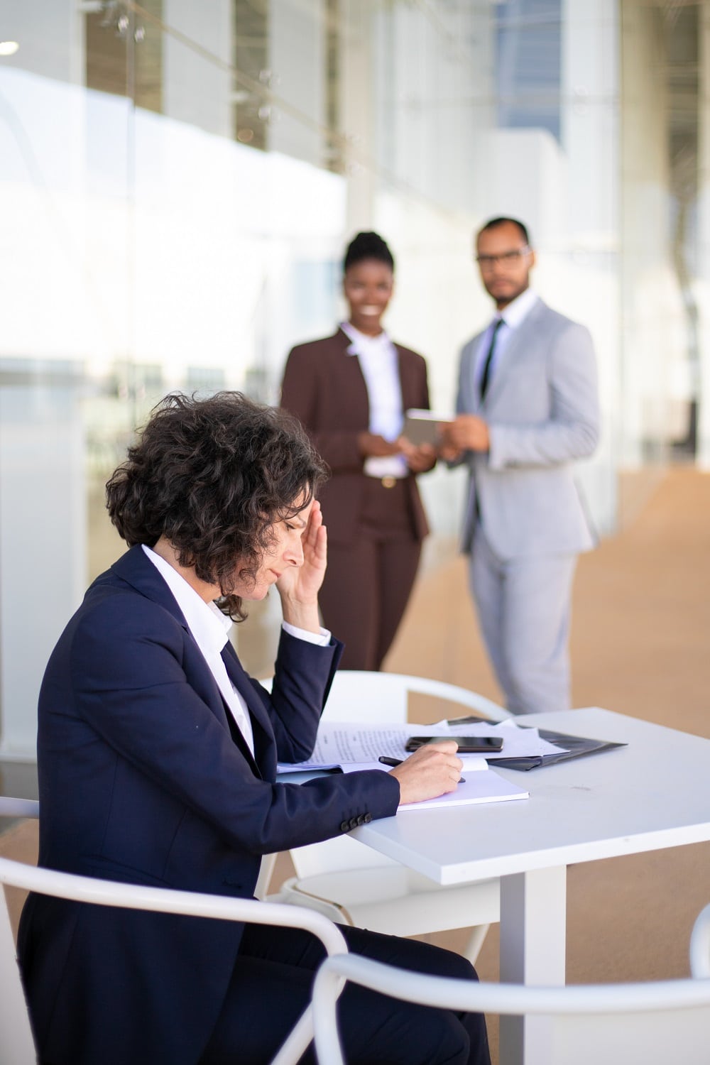 Frustrated tired female employee working with documents and checking noted. Multiethnic colleagues standing in background and laughing. Office conflict concept